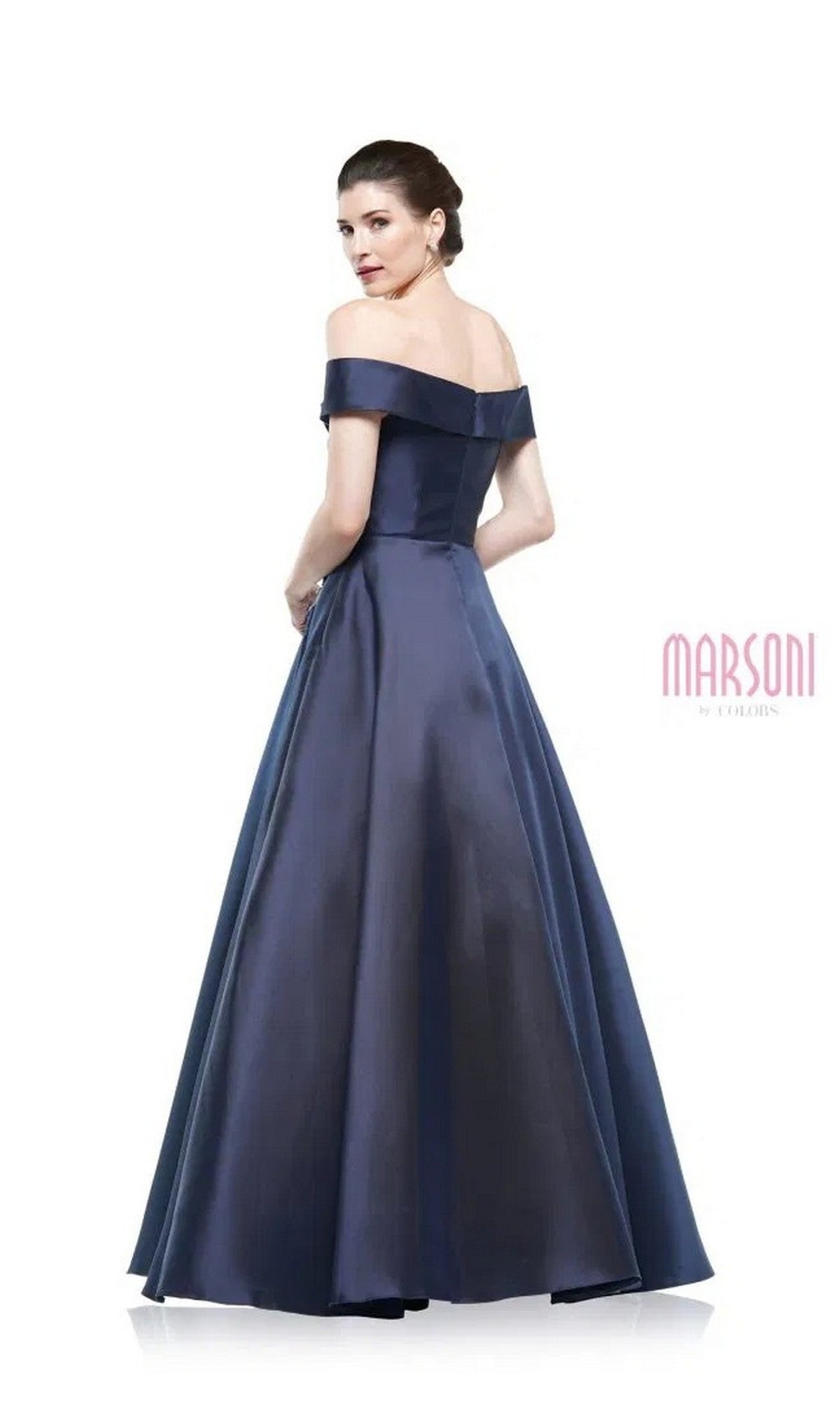 A woman wears the Marsoni MV1008 long formal dress in navy blue, an off-the-shoulder, floor-length A-line gown with beaded applique, standing with her back slightly turned and looking over her shoulder.